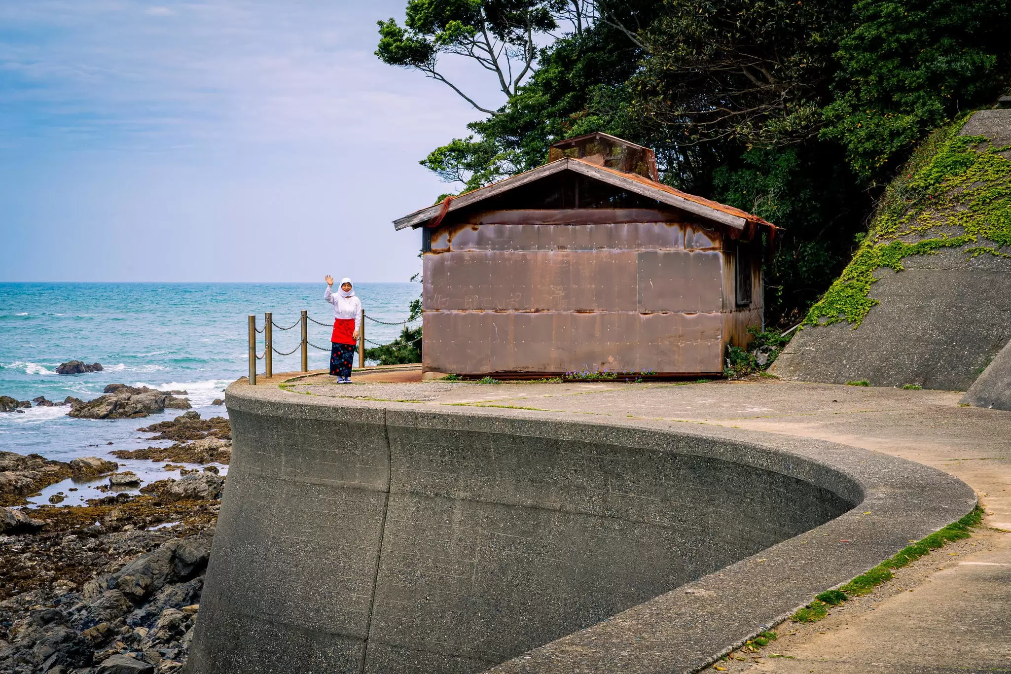 A woman in a long black skirt with a red apron, and a billowy white shirt and hood, waves; she stands beside a small hut beside the sea.