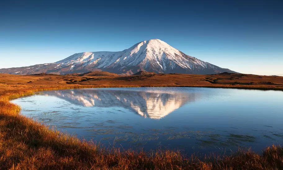 Snow-capped volcano reflected in a still tundra lake fringed by russet grass in Kamchatka, Russia