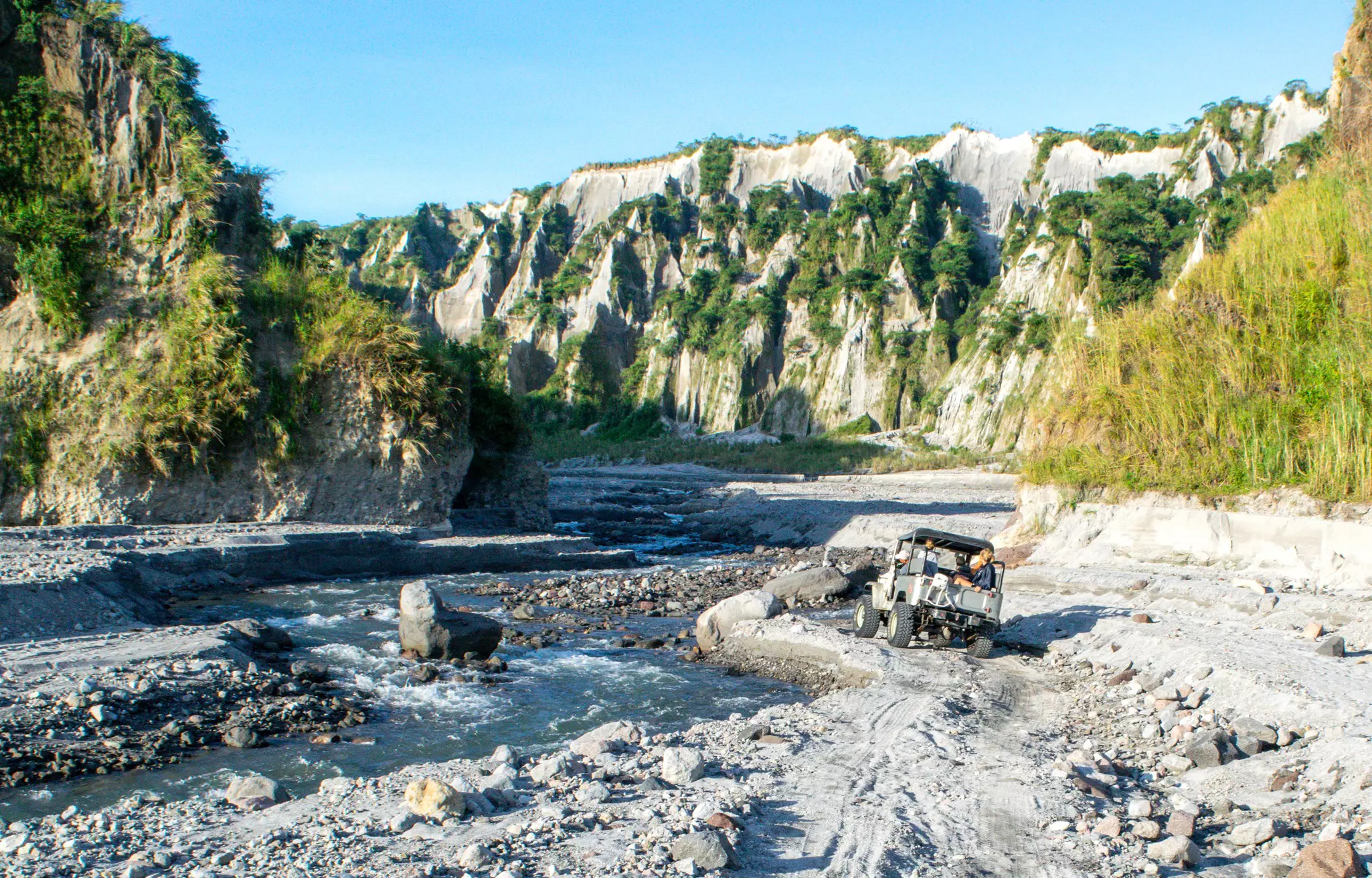 People ride in the back of a four-wheel drive on a tour of rocky landscape near a fast-flowing river.