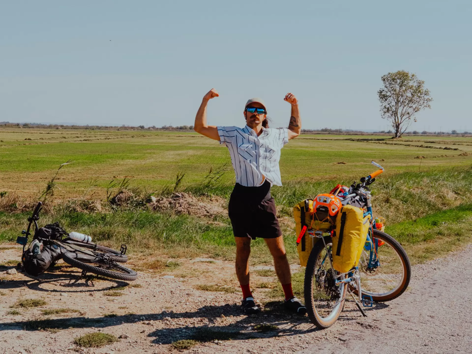 A man flexes his muscles on the side of the road next to two bikes. 