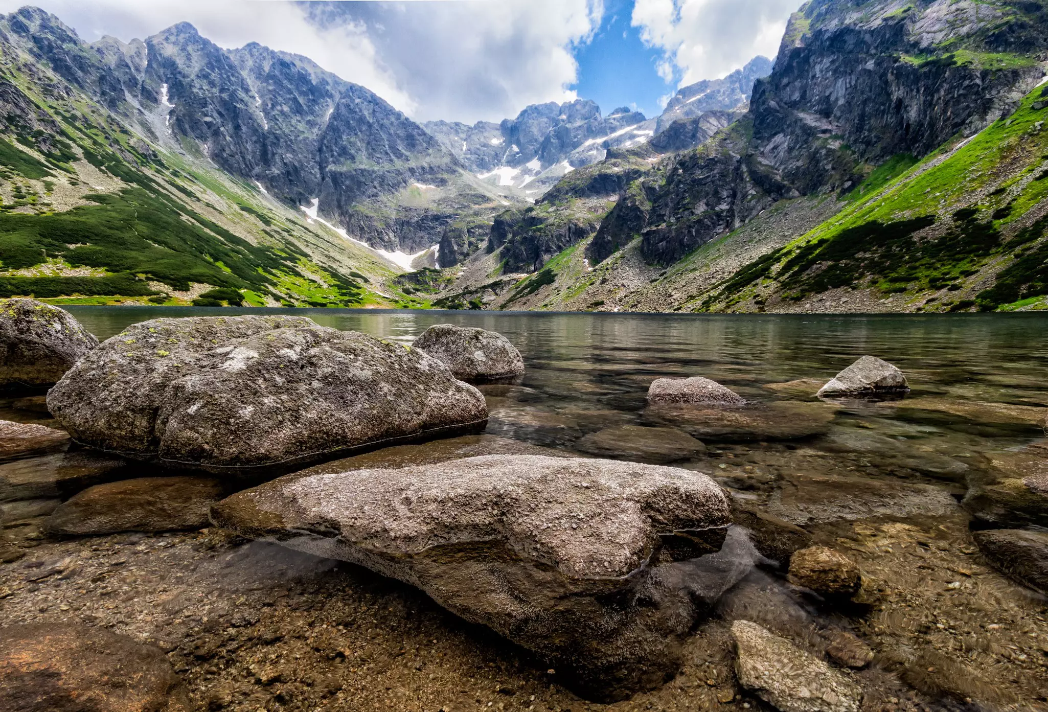 A clear mountain lake viewed from the perspective of the waterline; large stones are in the water, and mountains rise from the far shore.