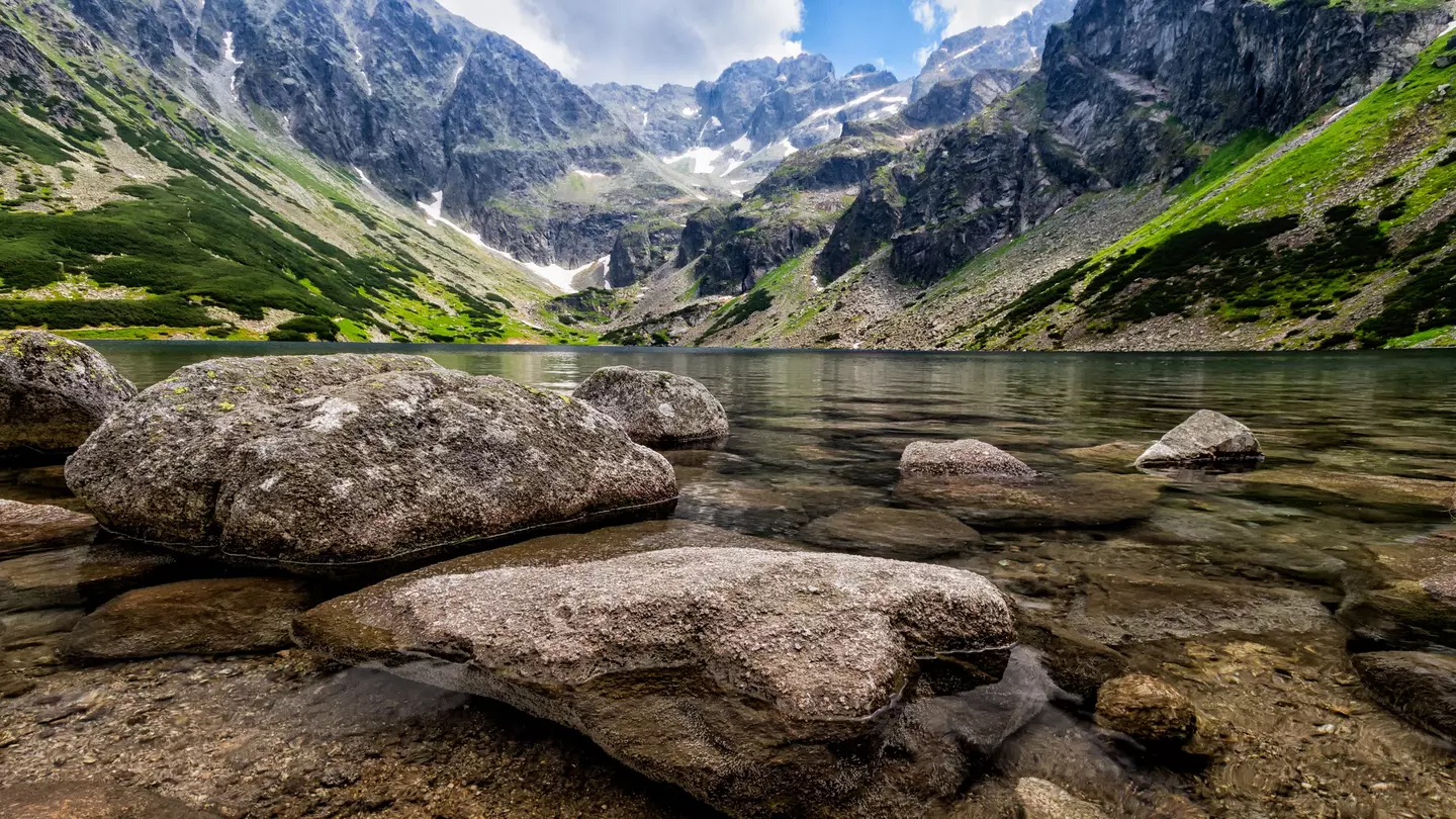 A clear mountain lake viewed from the perspective of the waterline; large stones are in the water, and mountains rise from the far shore.