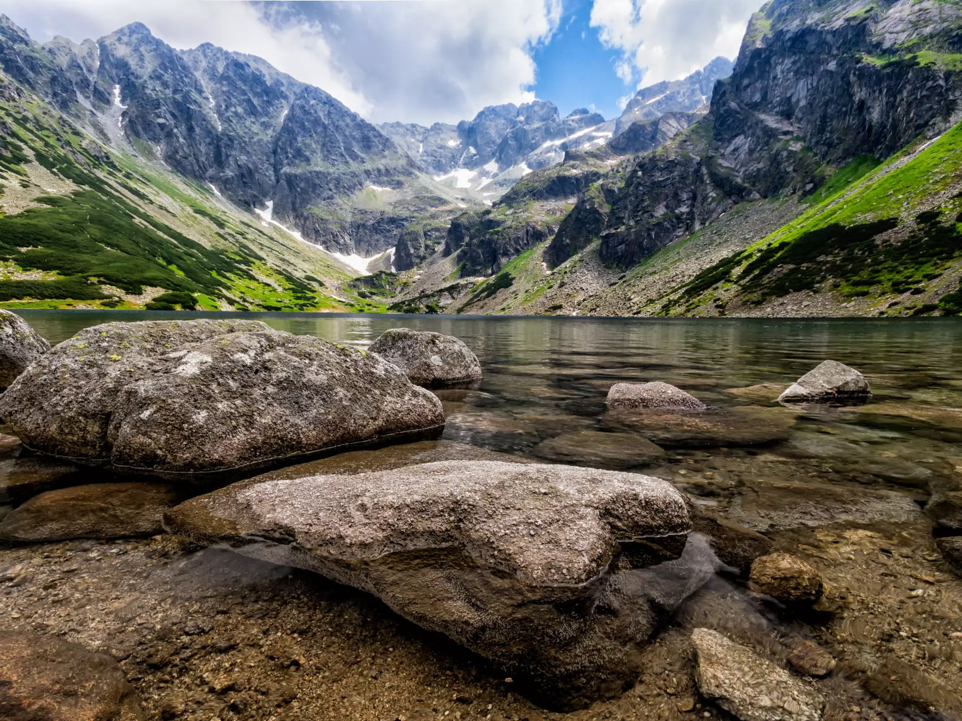 A clear mountain lake viewed from the perspective of the waterline; large stones are in the water, and mountains rise from the far shore.