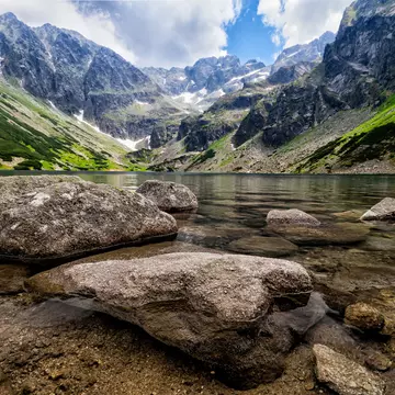 A clear mountain lake viewed from the perspective of the waterline; large stones are in the water, and mountains rise from the far shore.