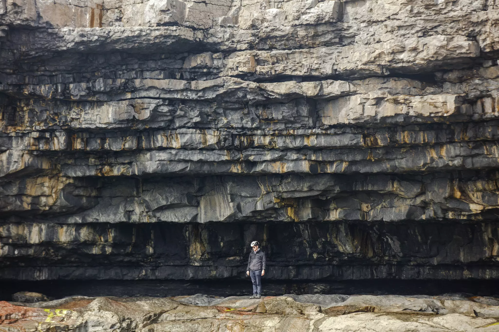 A man stands on an outcrop underneath bands of rock formations.