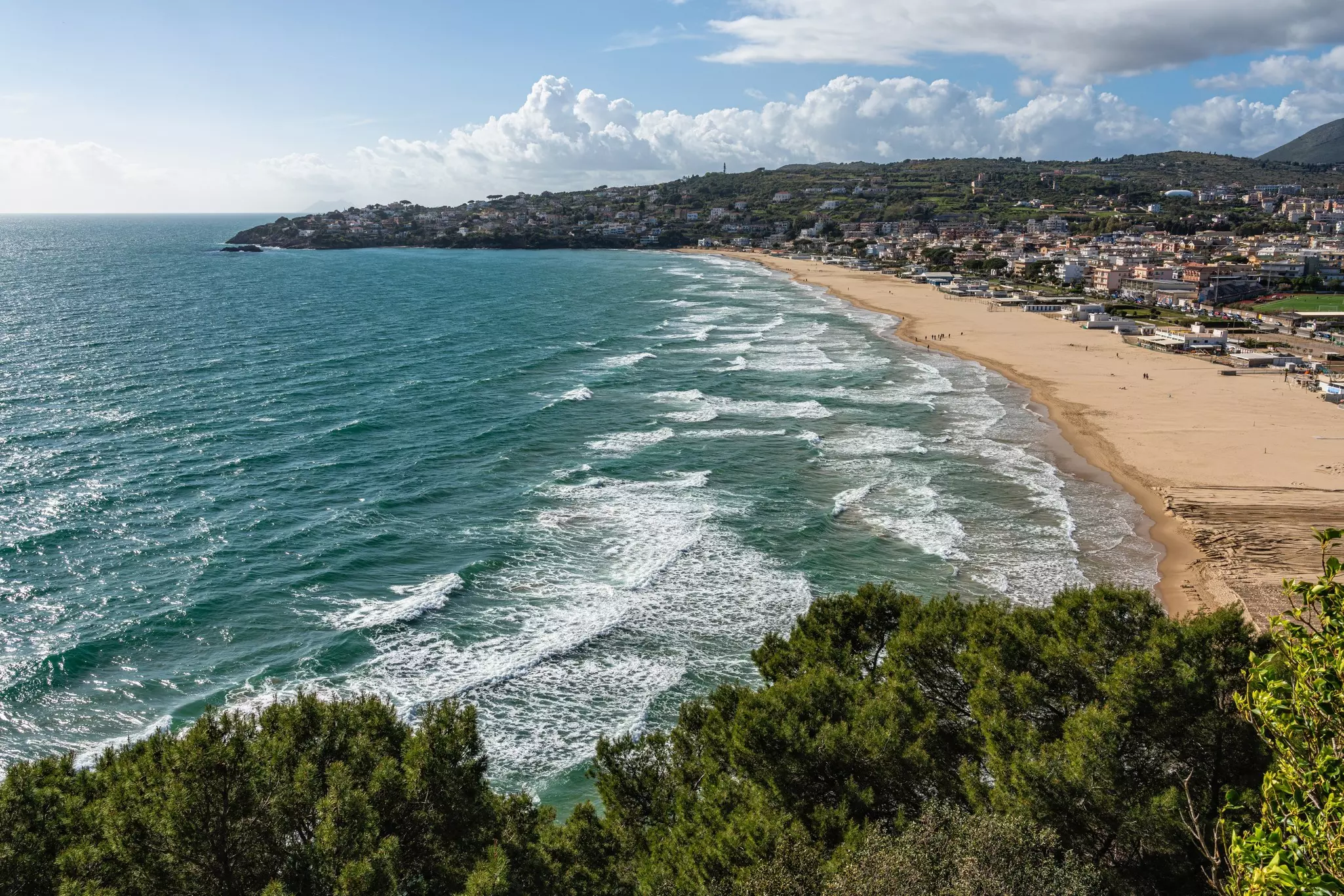 Aerial shot of ocean with sandy beach and buildings to the right and a curved peninsula and clouds in the distance.