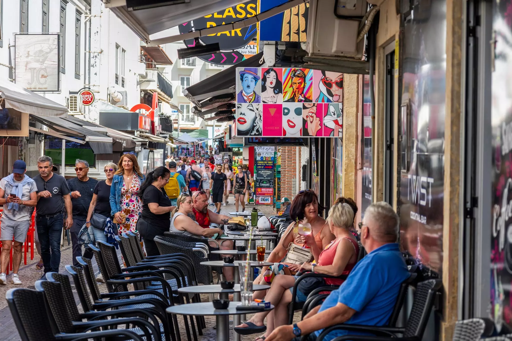 People sit at tables at an outdoor cafe on a busy pedestrianized street in a city.