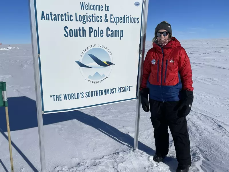 A woman stands beside a sign that says "South Pole Camp"