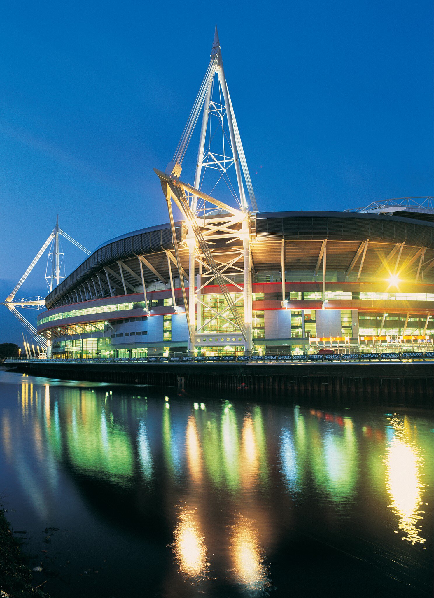 The Principality Stadium, formerly known as the Milllennium Stadium, in Cardiff, Wales. Night time with glowing lights reflecting on water.