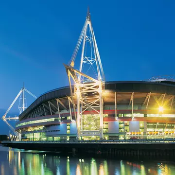 The Principality Stadium, formerly known as the Milllennium Stadium, in Cardiff, Wales. Night time with glowing lights reflecting on water.