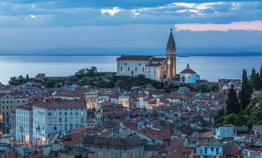 Piran's red-roofed old town at dusk with St George's Church and its lit bell tower above the Adriatic