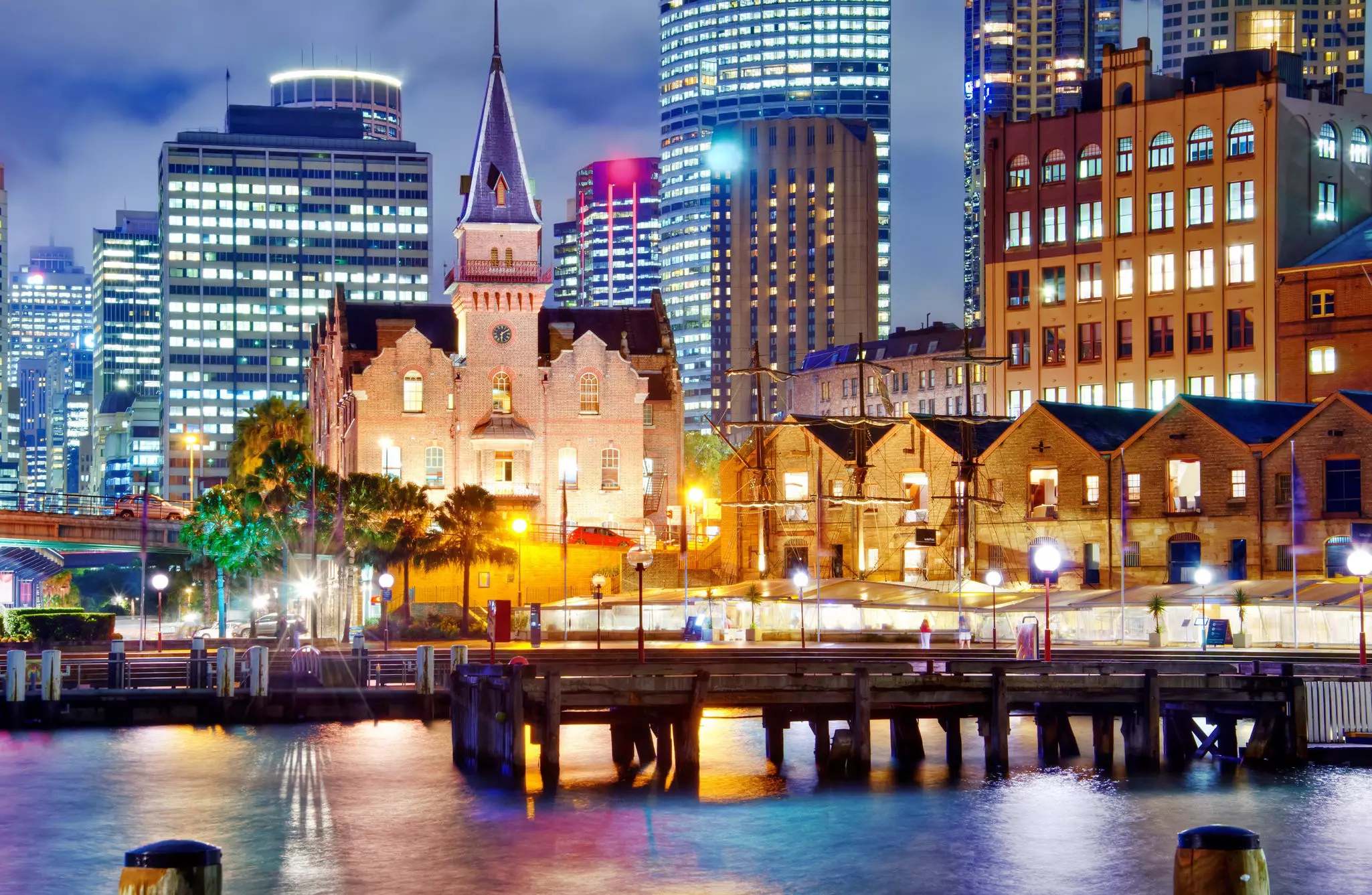 A city skyline at night with lights shining in building windows and from streetlights, with a harbor and pier in the foreground.