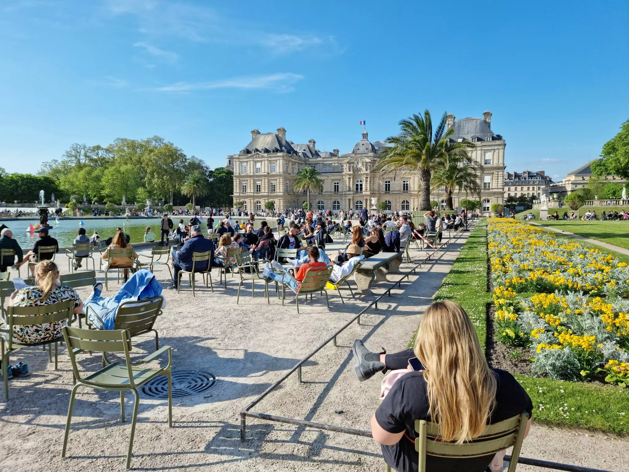 People relax in chairs and loungers on a stretch of gravel pathway set between flower-filled borders and a small artificial lake in a large public park overlooked by a grand mansion.
