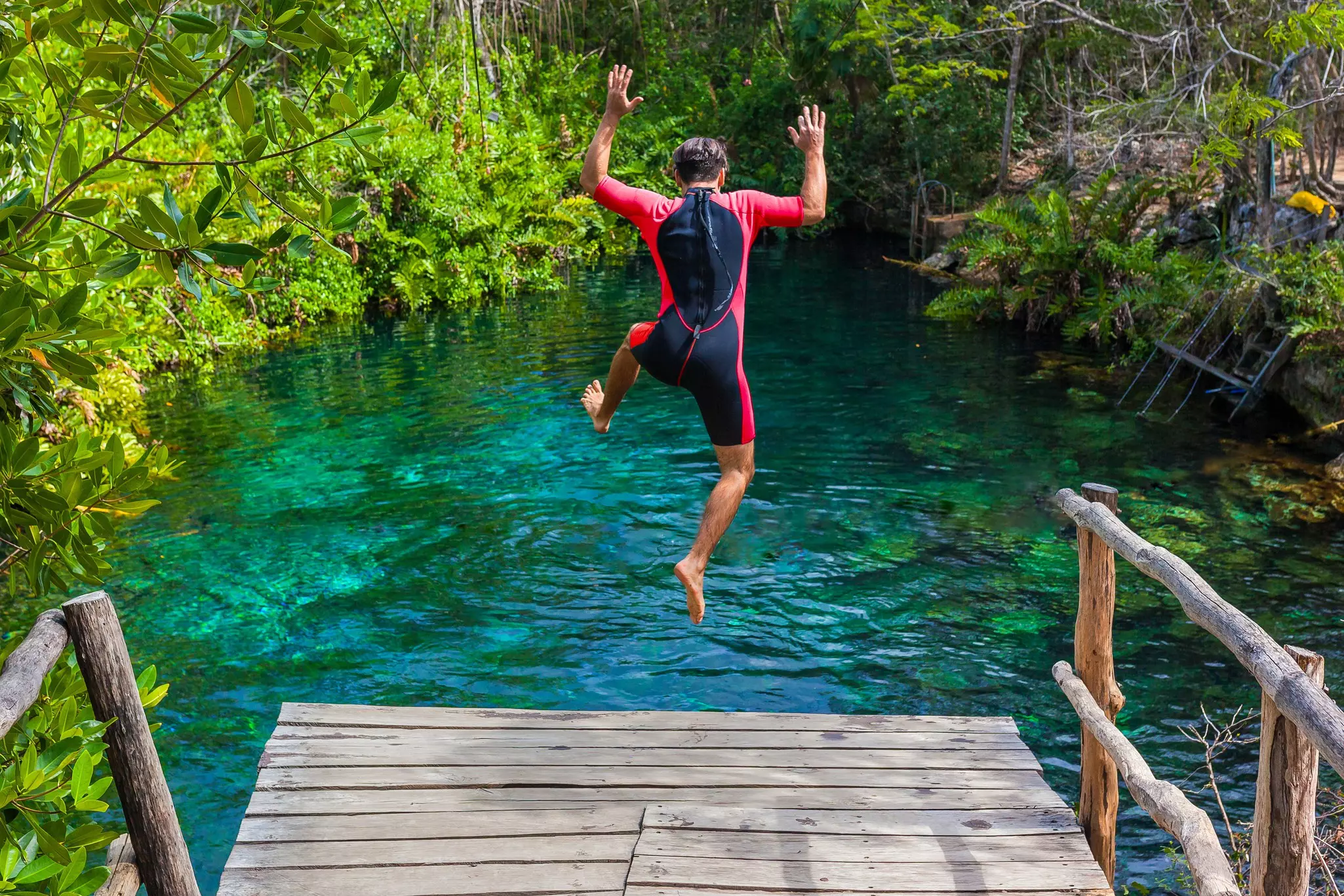 A man in a wetsuit jumps off a wooden platform into a freshwater lake surrounding by dense jungle.