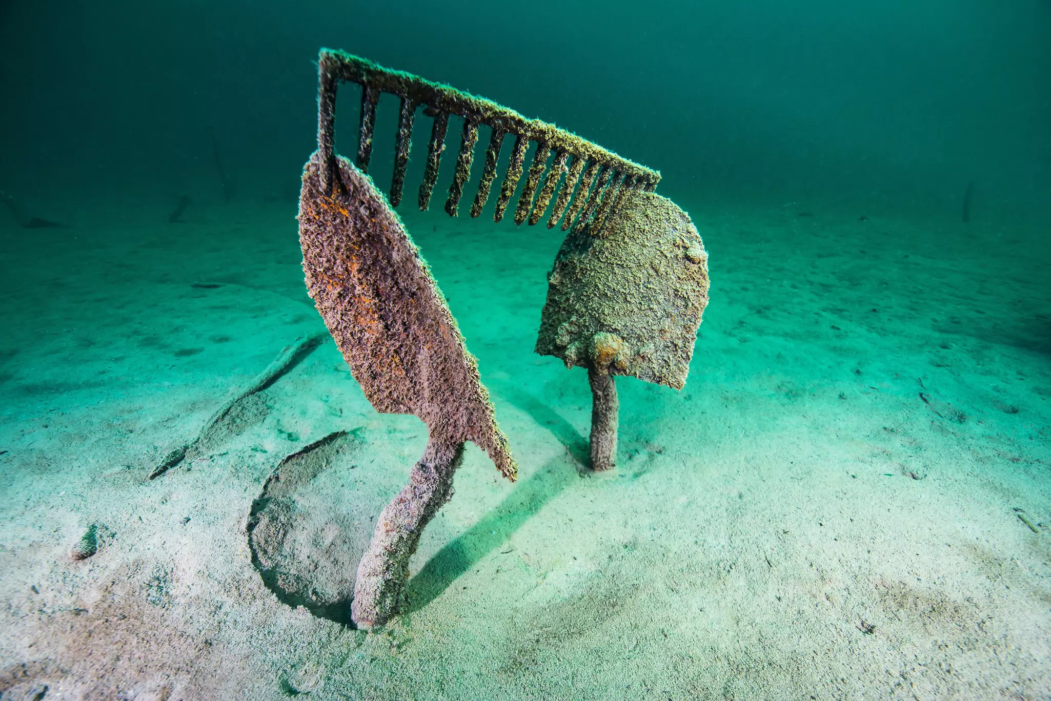 Two shovels, rusted and covered in marine sediment, stand straight up from the bed of Lake McDonald, with an equally rusted rake head perched on top. All around are the deep teal and bright aqua waters found throughout Glacier National Park