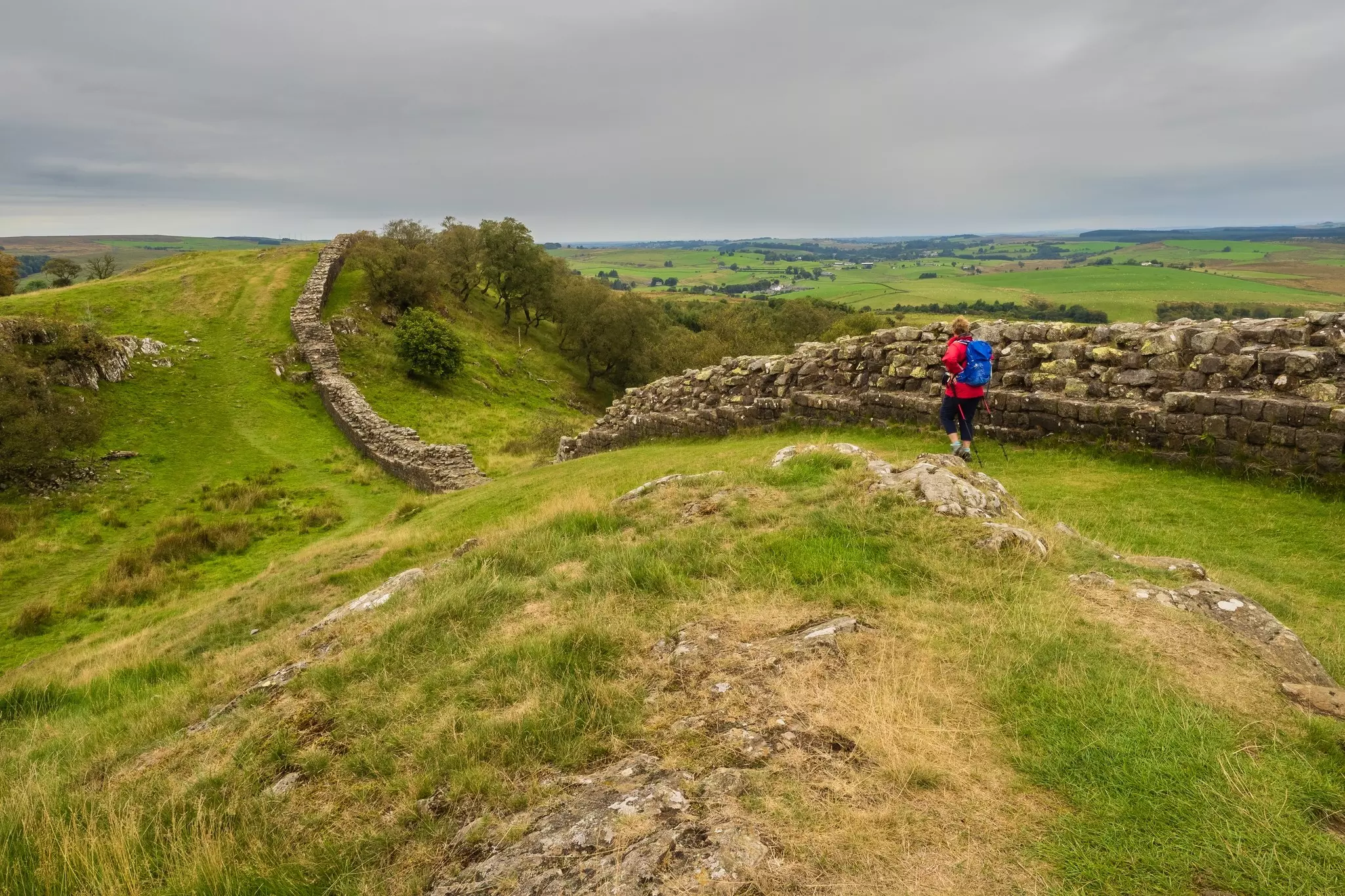 A hiker follows a path alongside the ruins of a Roman wall that runs over hillsides and into the distance in the countryside.