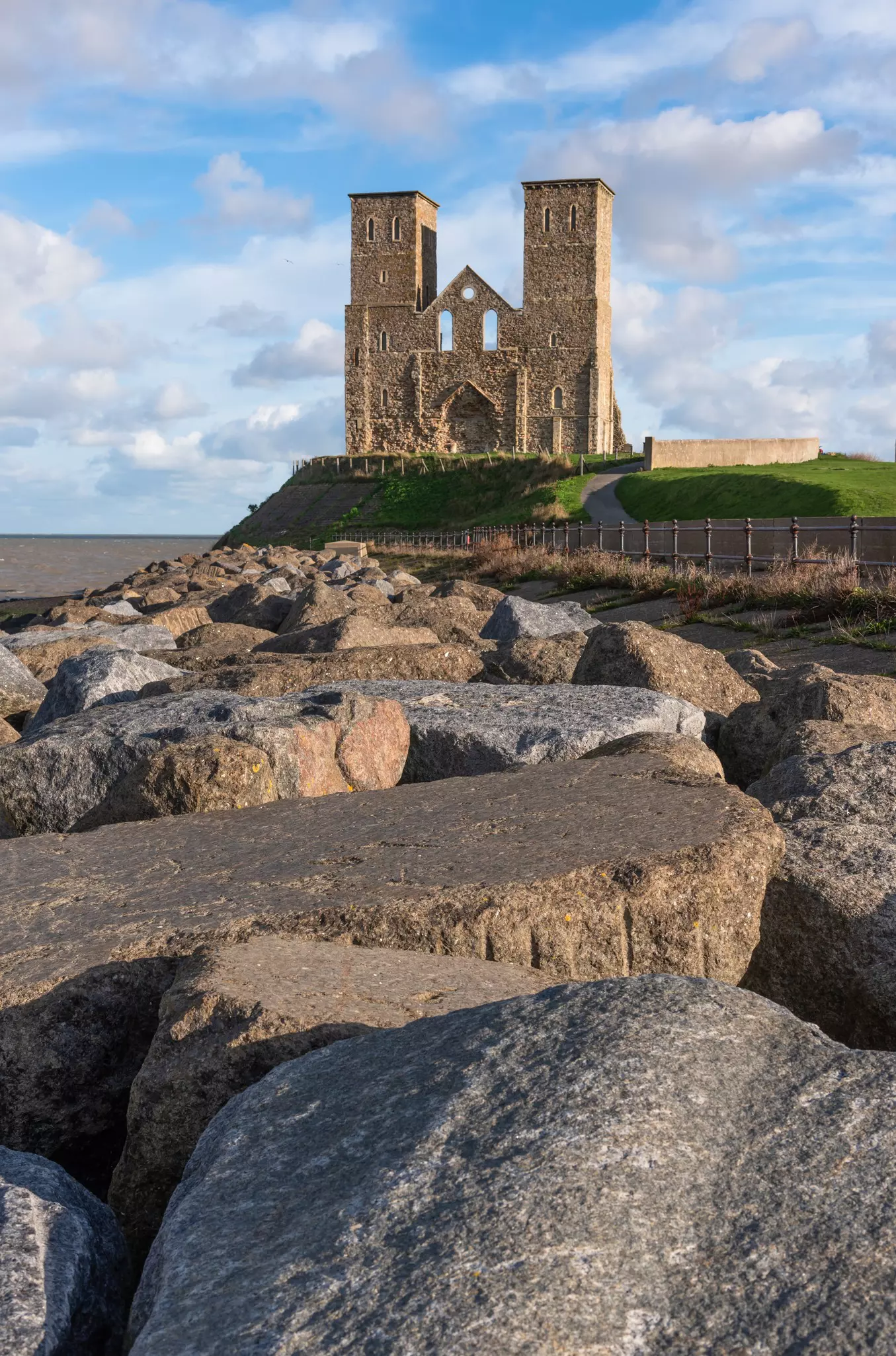 Large rocks are in the foreground; a cliff covered in green grass has the remains of a stone structure with towers on each side.