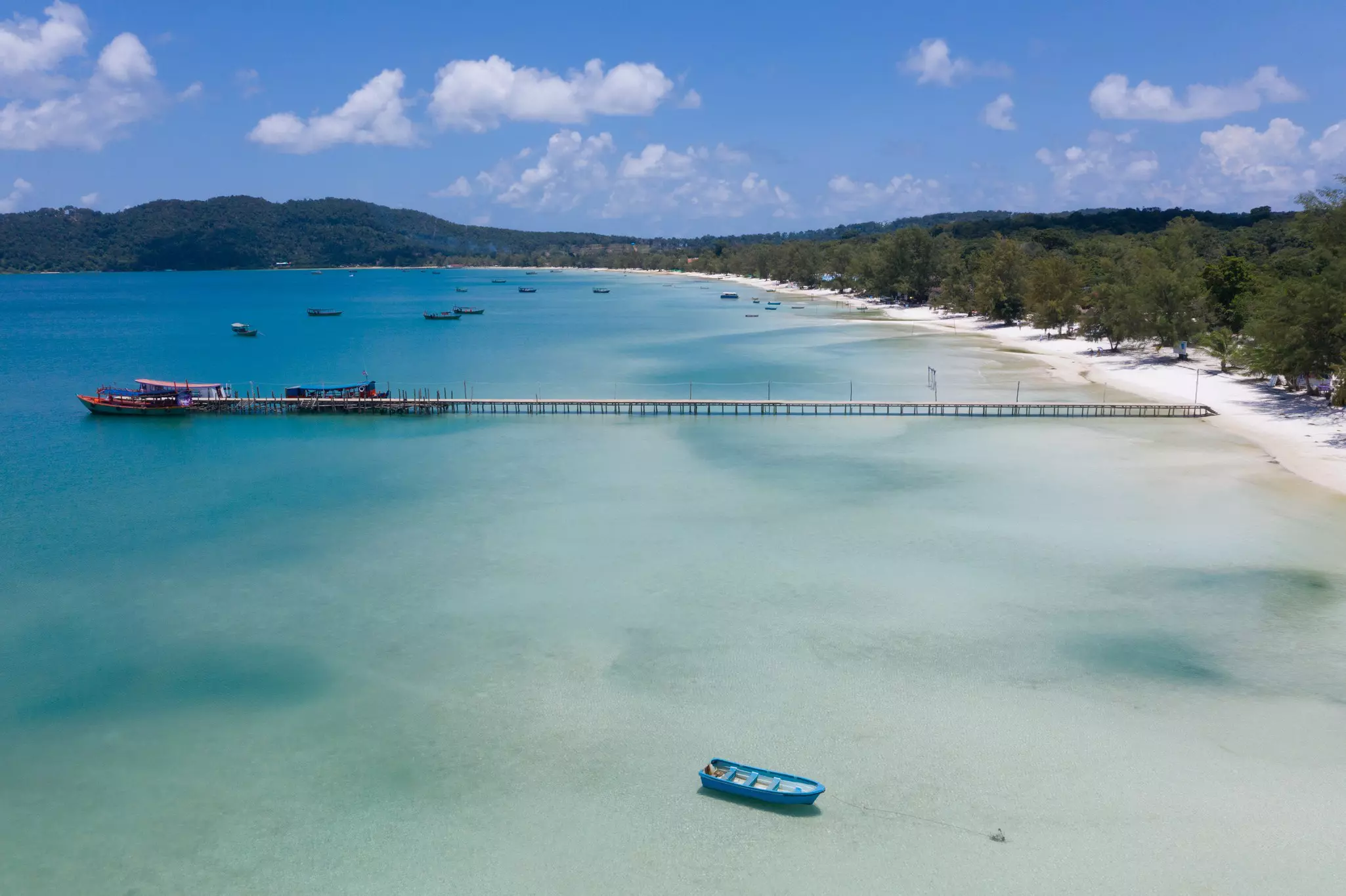 A long dock reaches from a white sand beach over blue water; a few boats are alongside the dock at the end.