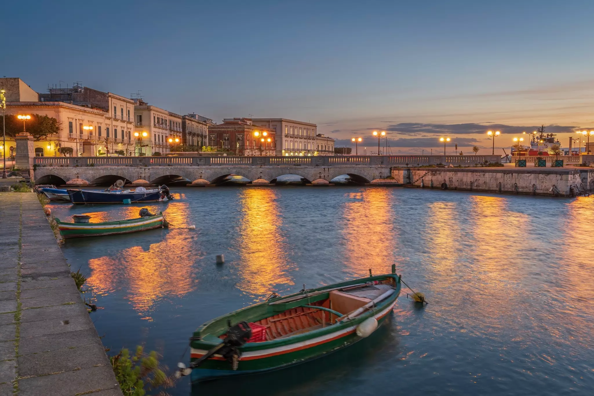 Sunset over a harbor in Italy; there are a few small boats in the water and a bridge with archways.