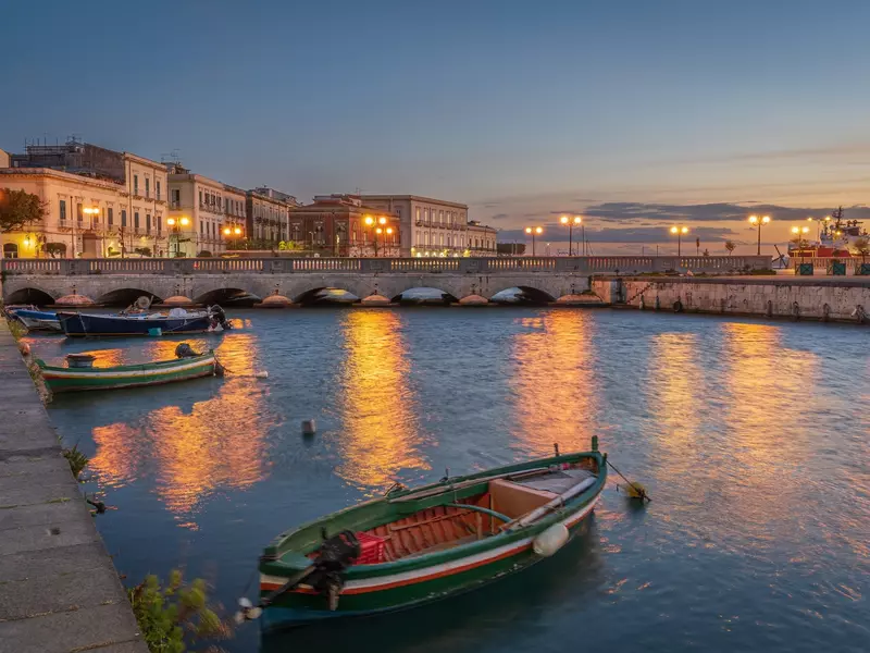 Sunset over a harbor in Italy; there are a few small boats in the water and a bridge with archways.