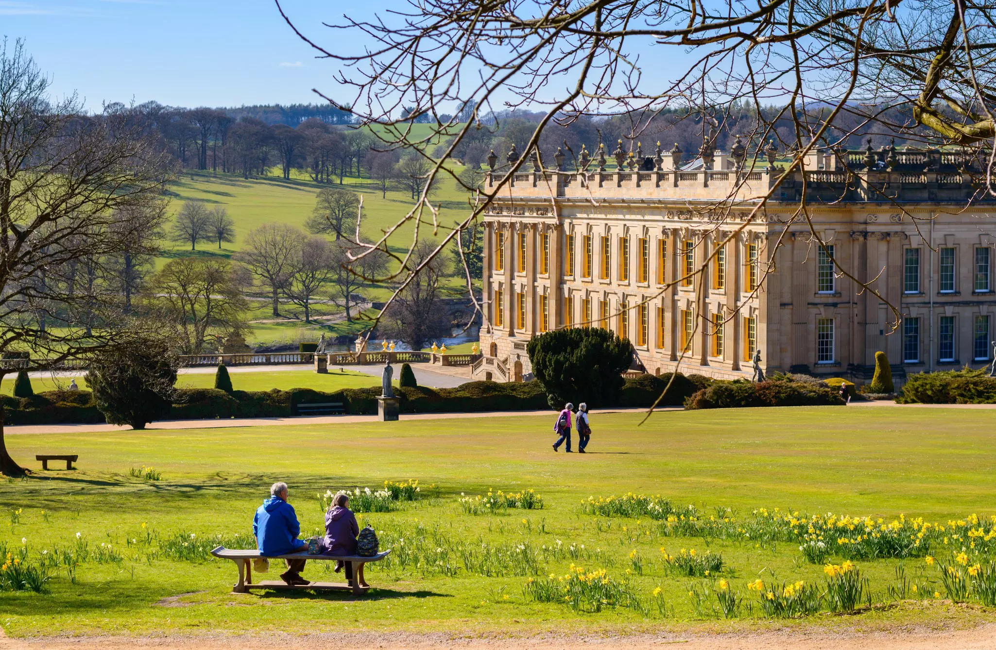 People sit on a bench on a lawn in front of an elaborate country mansion.