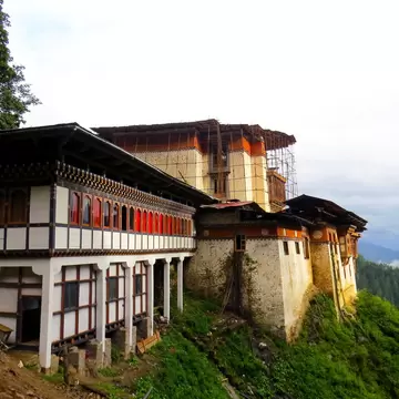 Whitewashed Tango Goemba monastery buildings with red-framed windows on a forested hillside, mountains beyond.