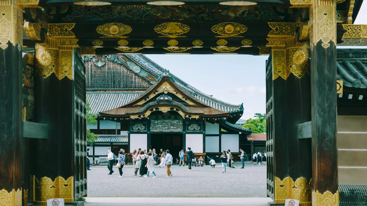 People are seen through the gate of a castle congregating in an inner courtyard. Elaborate gold decorations cover the gate and eaves of the temple beyond the courtyard.
