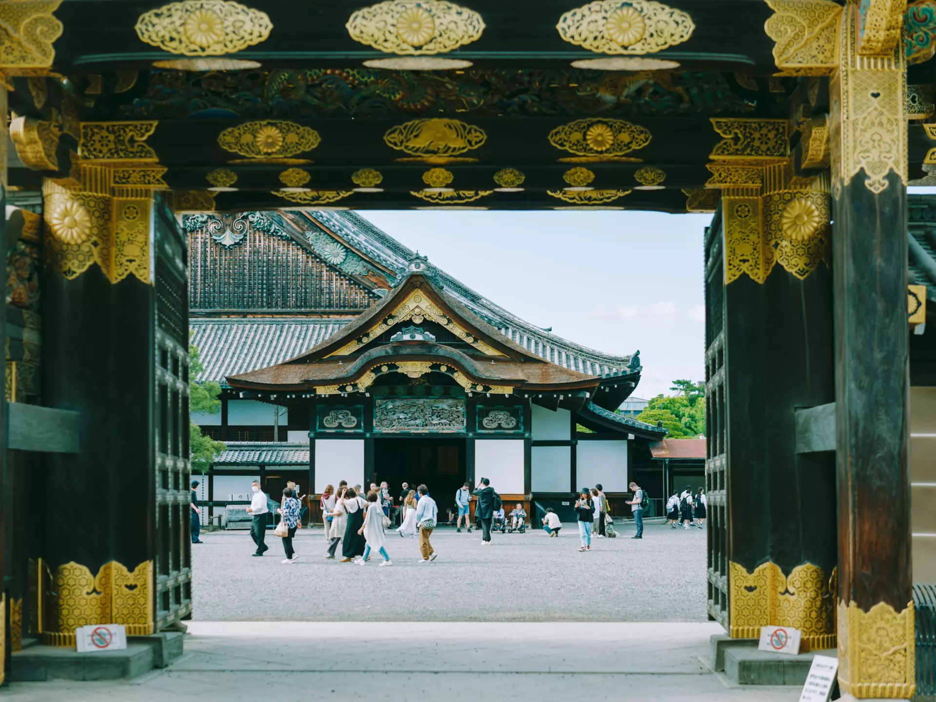 People are seen through the gate of a castle congregating in an inner courtyard. Elaborate gold decorations cover the gate and eaves of the temple beyond the courtyard.