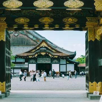 People are seen through the gate of a castle congregating in an inner courtyard. Elaborate gold decorations cover the gate and eaves of the temple beyond the courtyard.