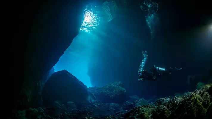 Two people scuba diving in a cave, with light shining through a hole in the cave