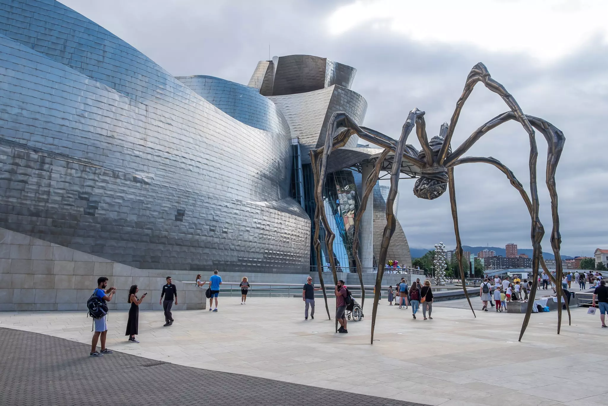People walk past a large sculpture of a spider on a terrace outside a museum building covered in titanium tiles.