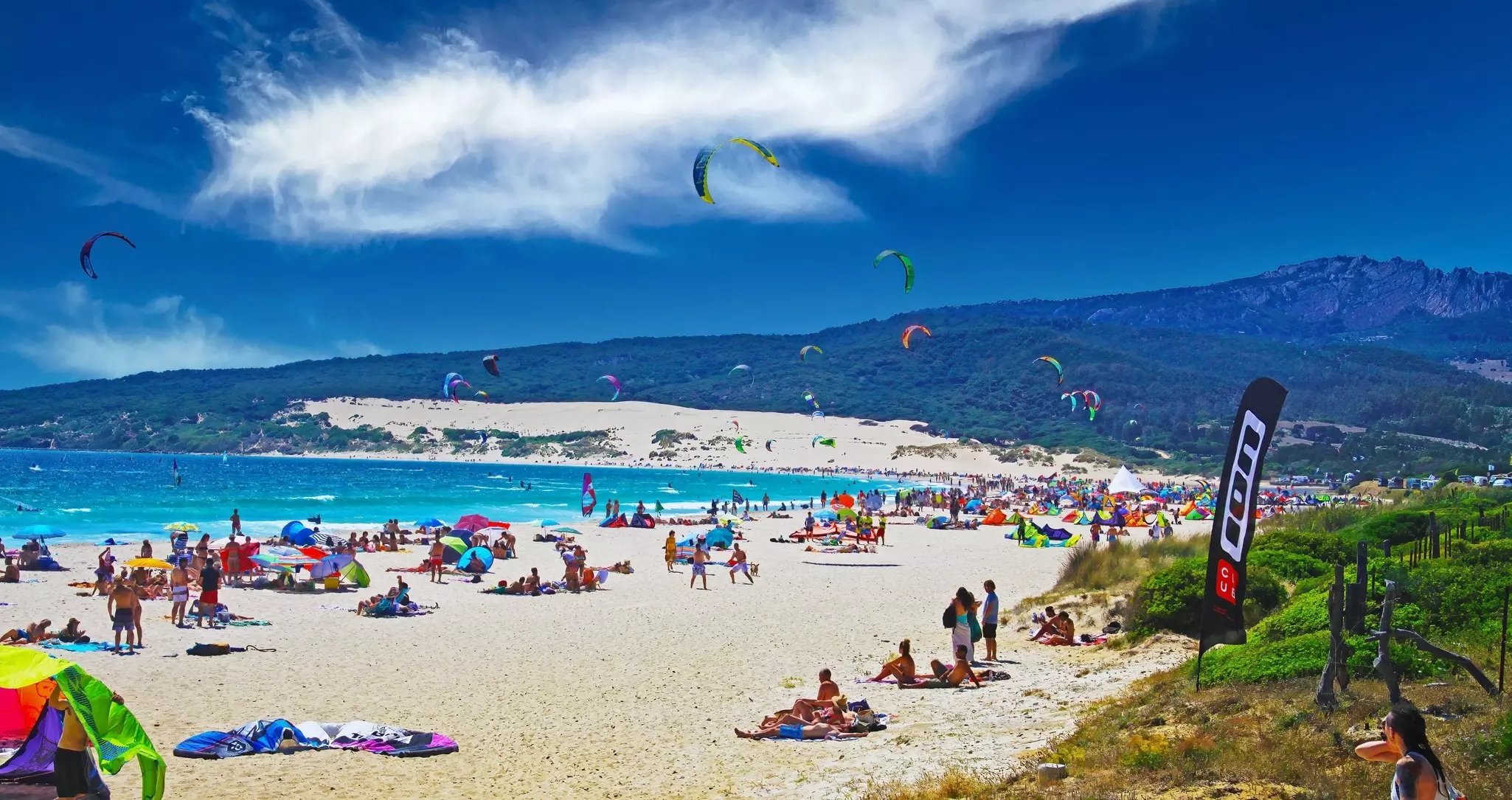 Beautiful natural crowded lagoon sand beach, kite surfers kitesurfing, dunes, green hill, blue sky at Playa Bolonia. 
