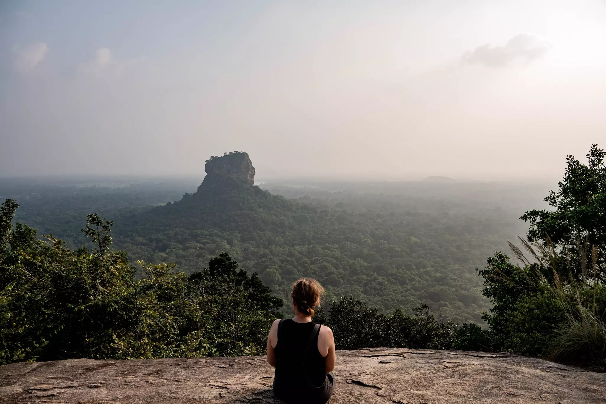 A woman sits on the rock of Pidurangala looking towards another large jungle-covered rocky outcrop in Sigiriya, Sri Lanka