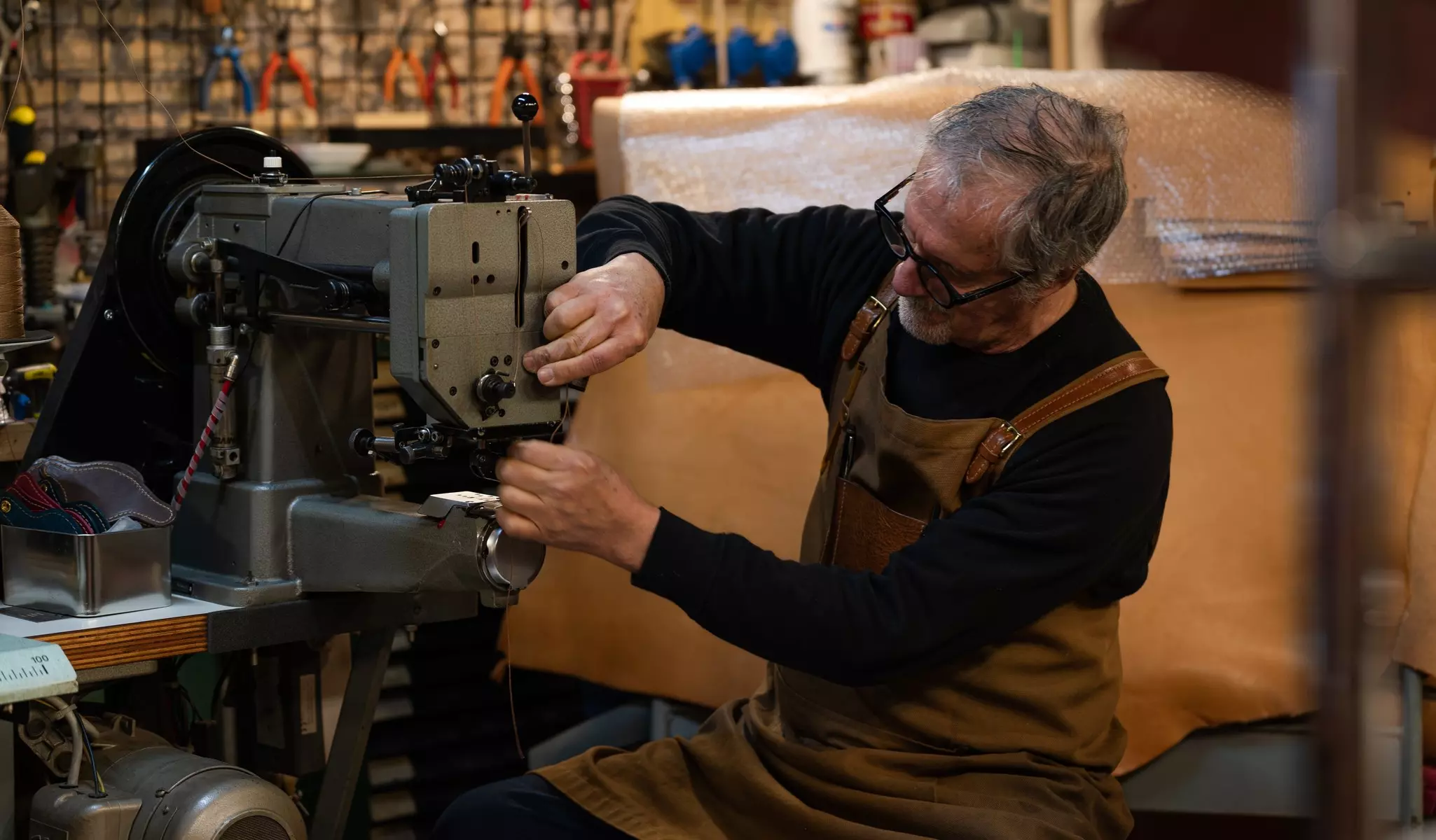 adult craftsman sitting adding thread in a sewing machine to start sewing parts of a chair made of leather in his workshop, downtown rome, italy