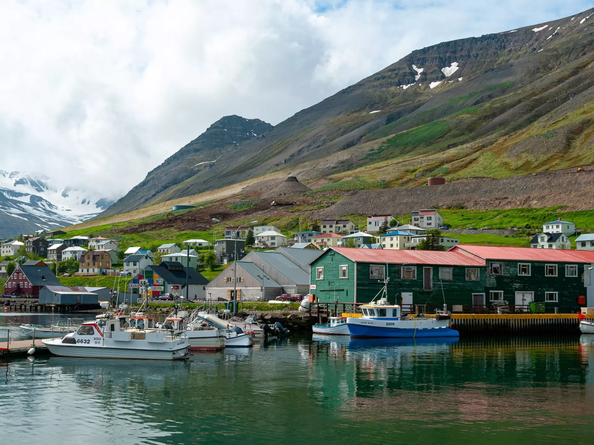 Siglufjörður harbor in North Iceland with fishing boats, colorful houses and church surrounded by mountains. Scenic coastal town with calm water and traditional village atmosphere in Icelandic landscape.