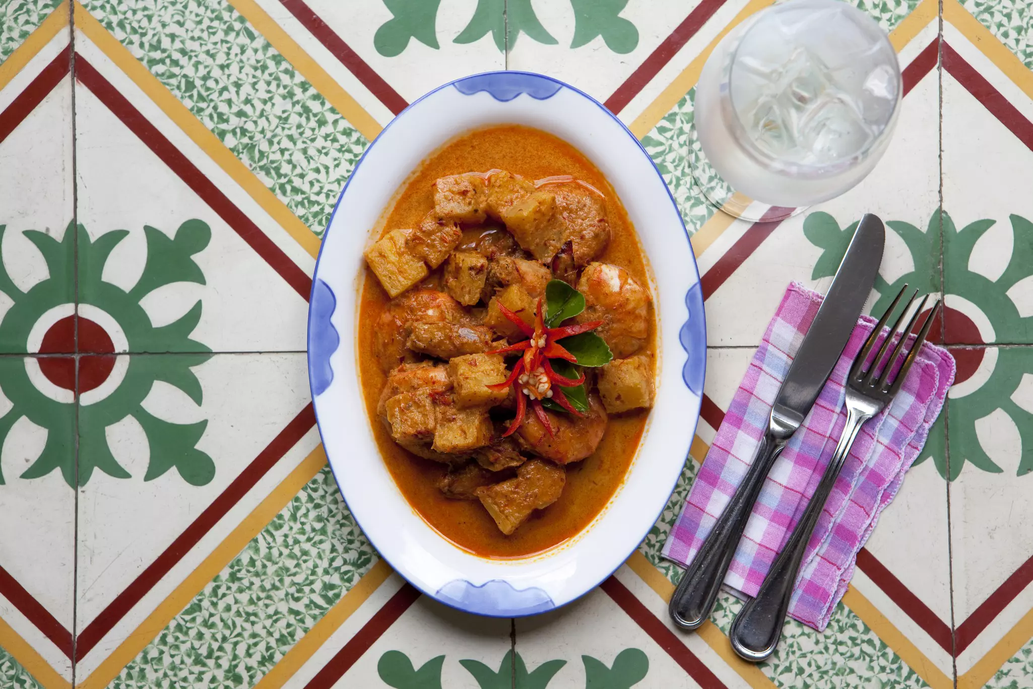 An overhead shot of a curry dish laid out on a tiled table, next to a fork and knife on a plaid napkin.
