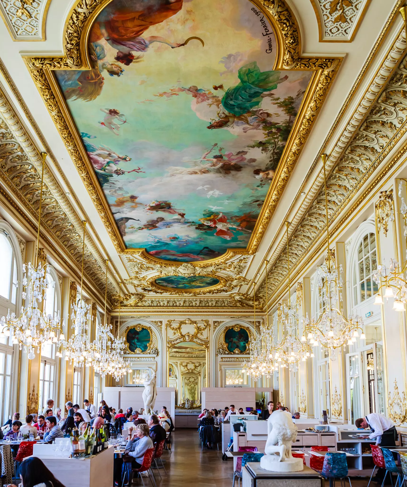 People eat at tables in a dining room with elaborate gilt decoration and a painted ceiling.