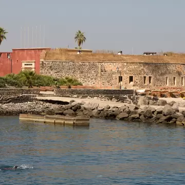 This image looks across the water to Île de Gorée and a circular stone fortress, with its small windows. To the left of the fortress is a pinkish section of the fortress, with palms in the background. A boat floats in the foreground.