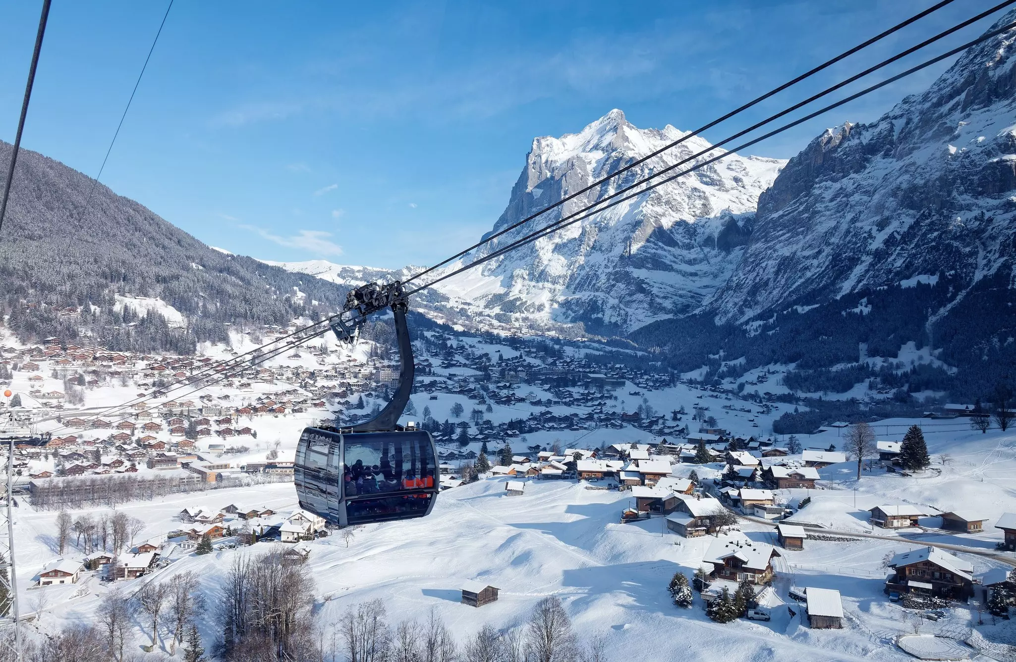 A cable car ascends over a snow-covered town and valley, with peaks visible in the distance.