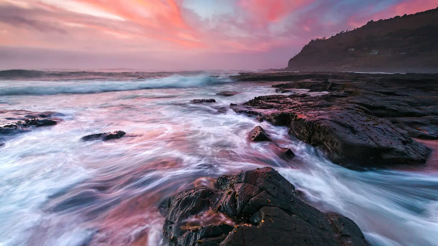 Waves wash onto coastal rocks with colourful clouds in the sky, Garie Beach.
