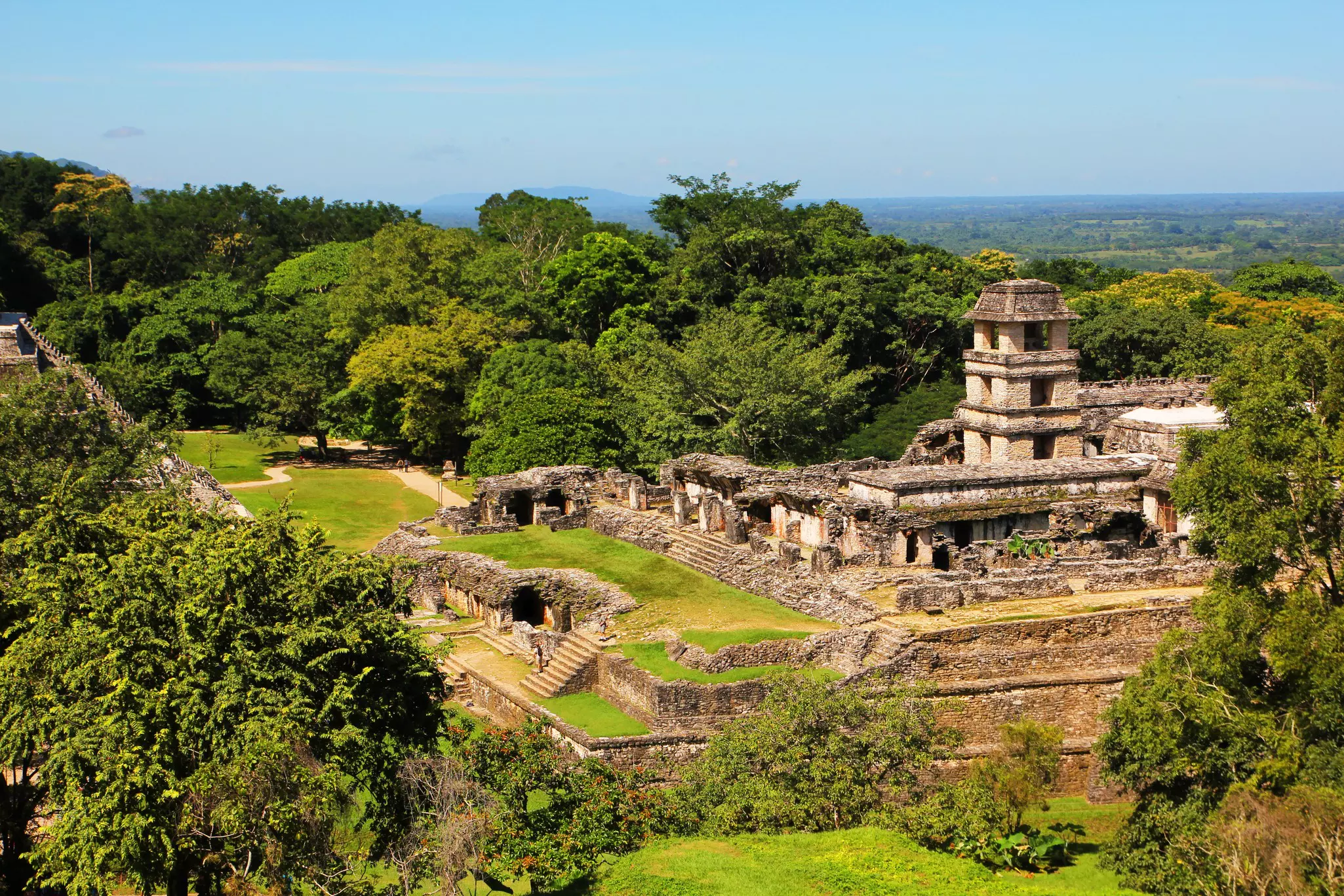 Aerial shot of Mayan ruins made of stone -- a tall tower and walls surrounded by green grasa and trees with a hazy blue sky in the background.