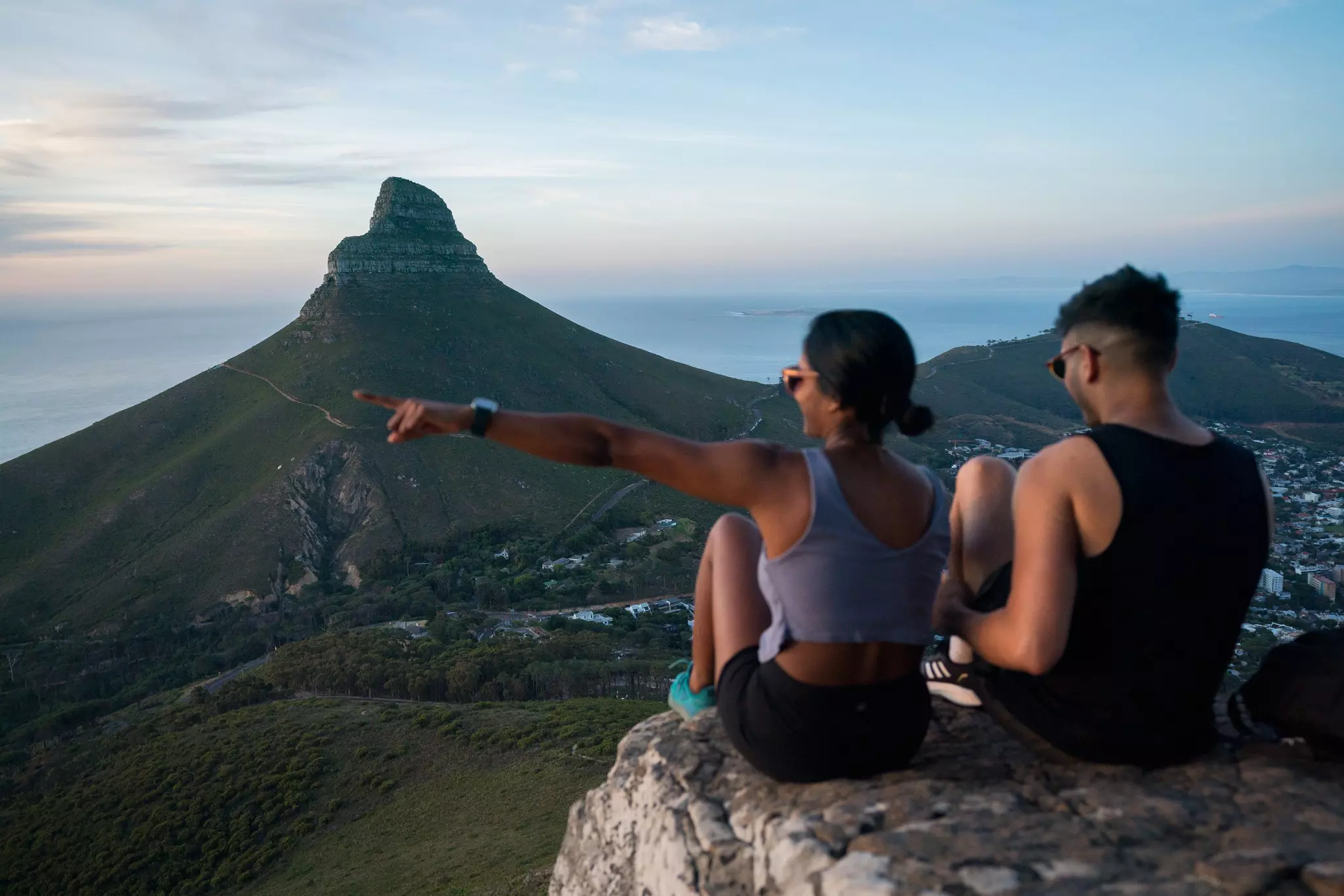 Two people are seen from behind sitting on a ledge overlooking a valley at dusk. A the pointed summit of a rocky outcrop is seen in the distance.