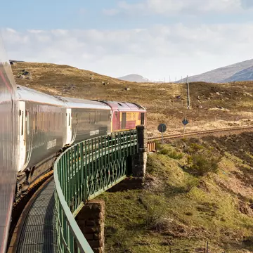 A silver train rumbles along a section of track with wide green fields located on either side of the track and low mountains in the distance on a sunny day.