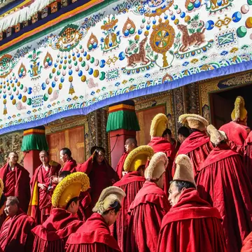 A crowd of monks in crimson robes and tall yellow hats filing beneath an ornate painted canopy into a monastery hall.