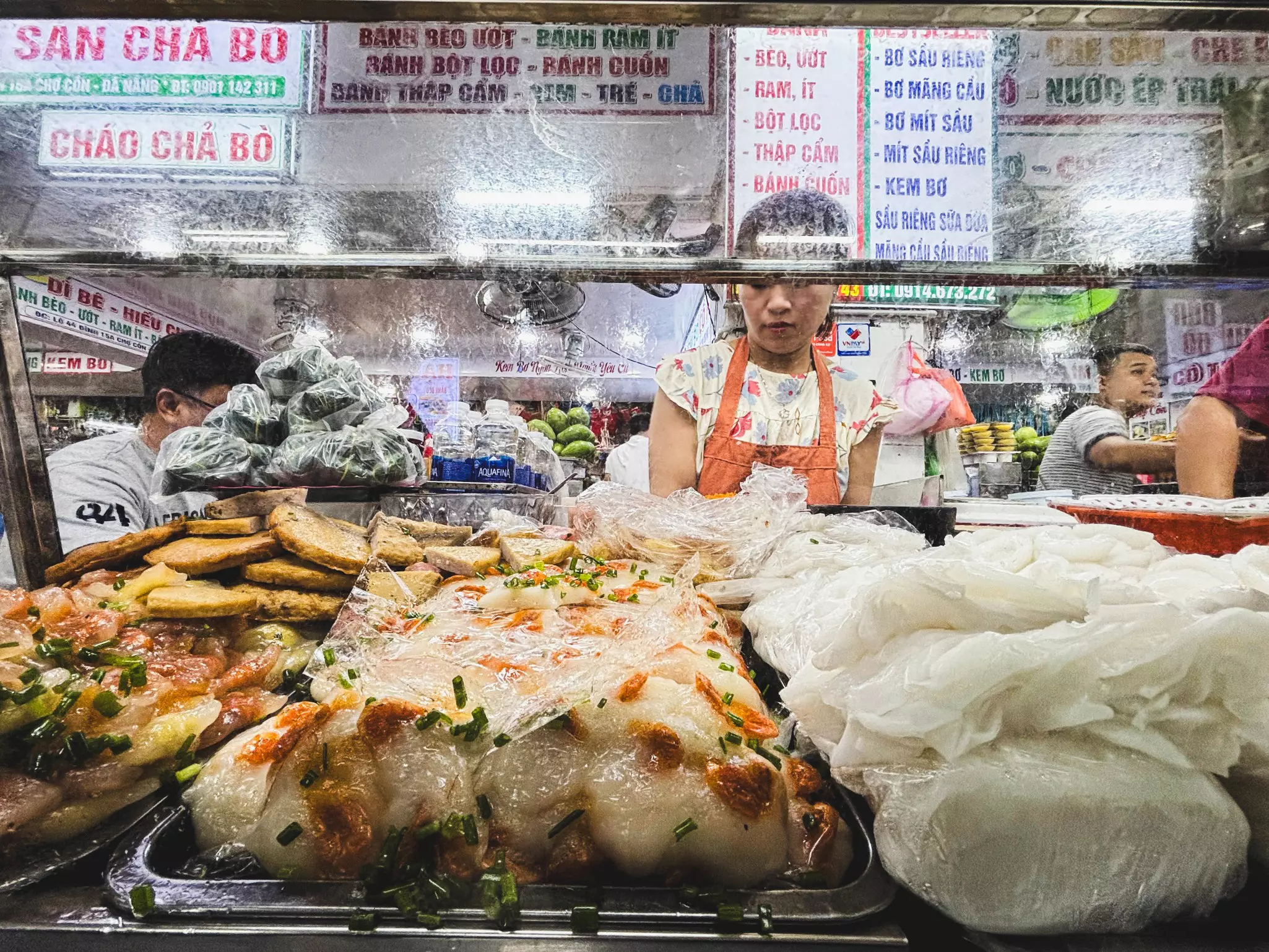 An assortment of rice flour cakes arranged on trays at a market. A woman in an orange apron stands behind the display