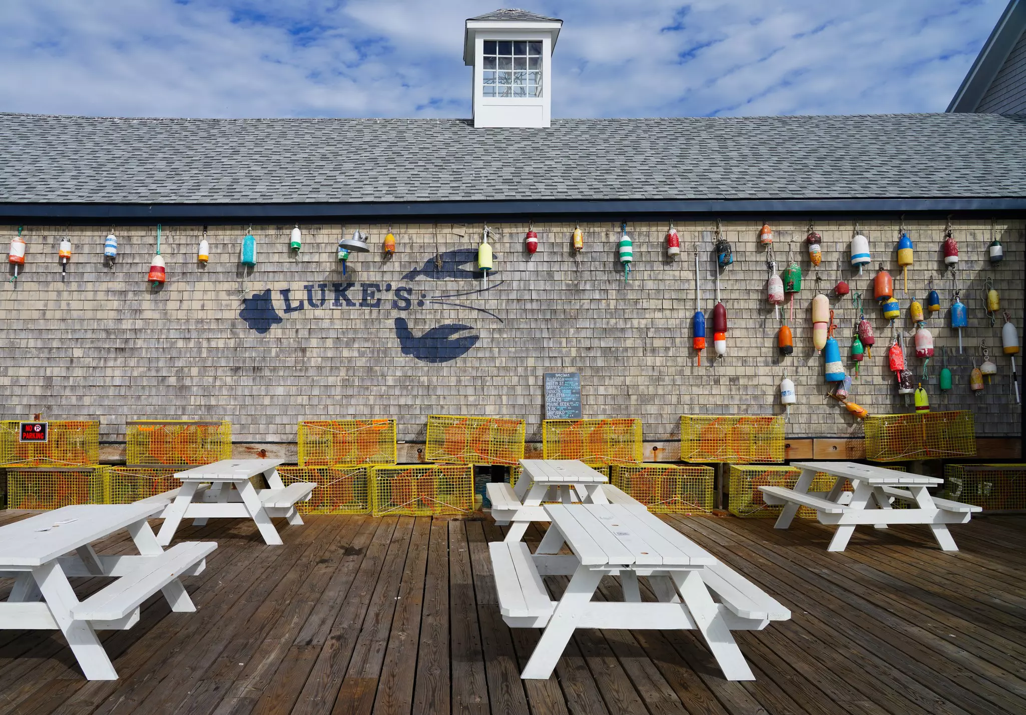 Side of a shingled building with Luke's painted on the side and wooden fishing buoys hanging. Yellow lobster cages are stacked against the wall, with white picnic benches in the foreground