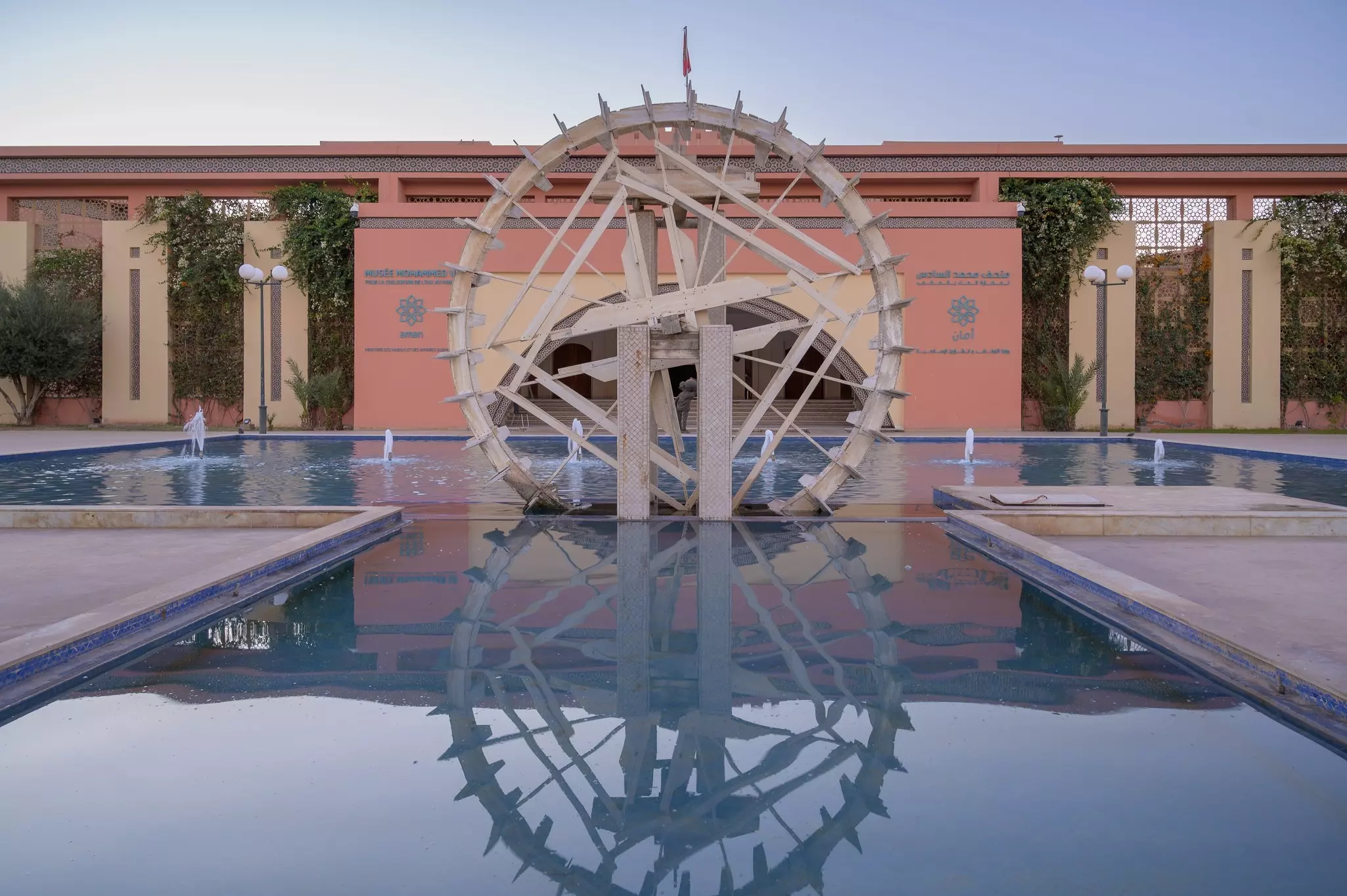 A large water wheel sits in a pool of water with a row of fountains outside a museum building.