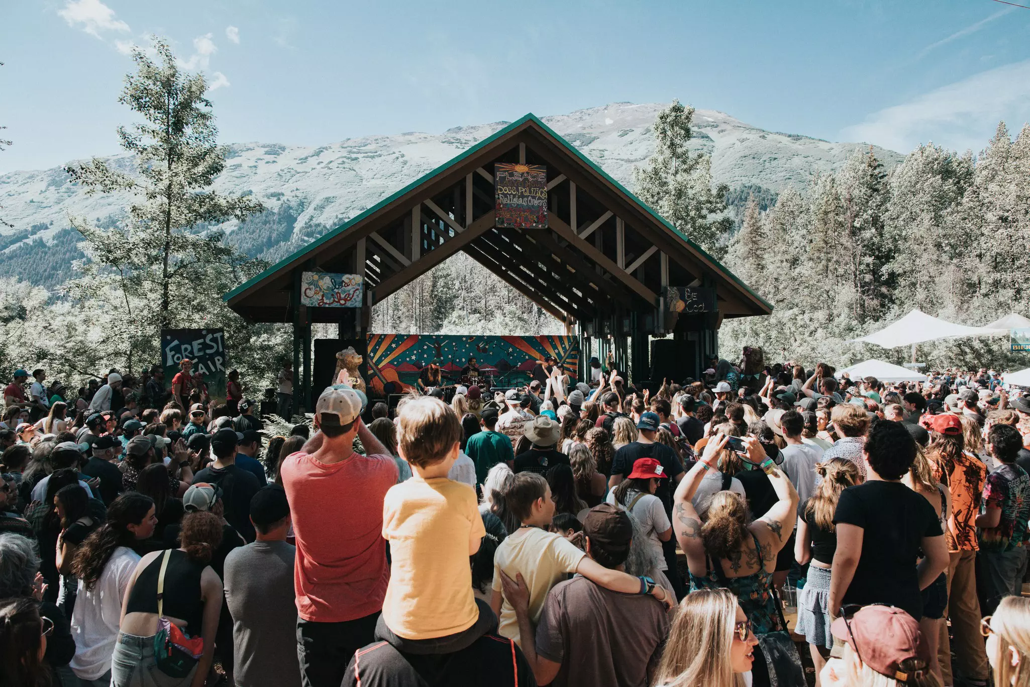 A crowd of people gathers in front of a wooden stage while musicians play. Mountains and evergreens in the background.