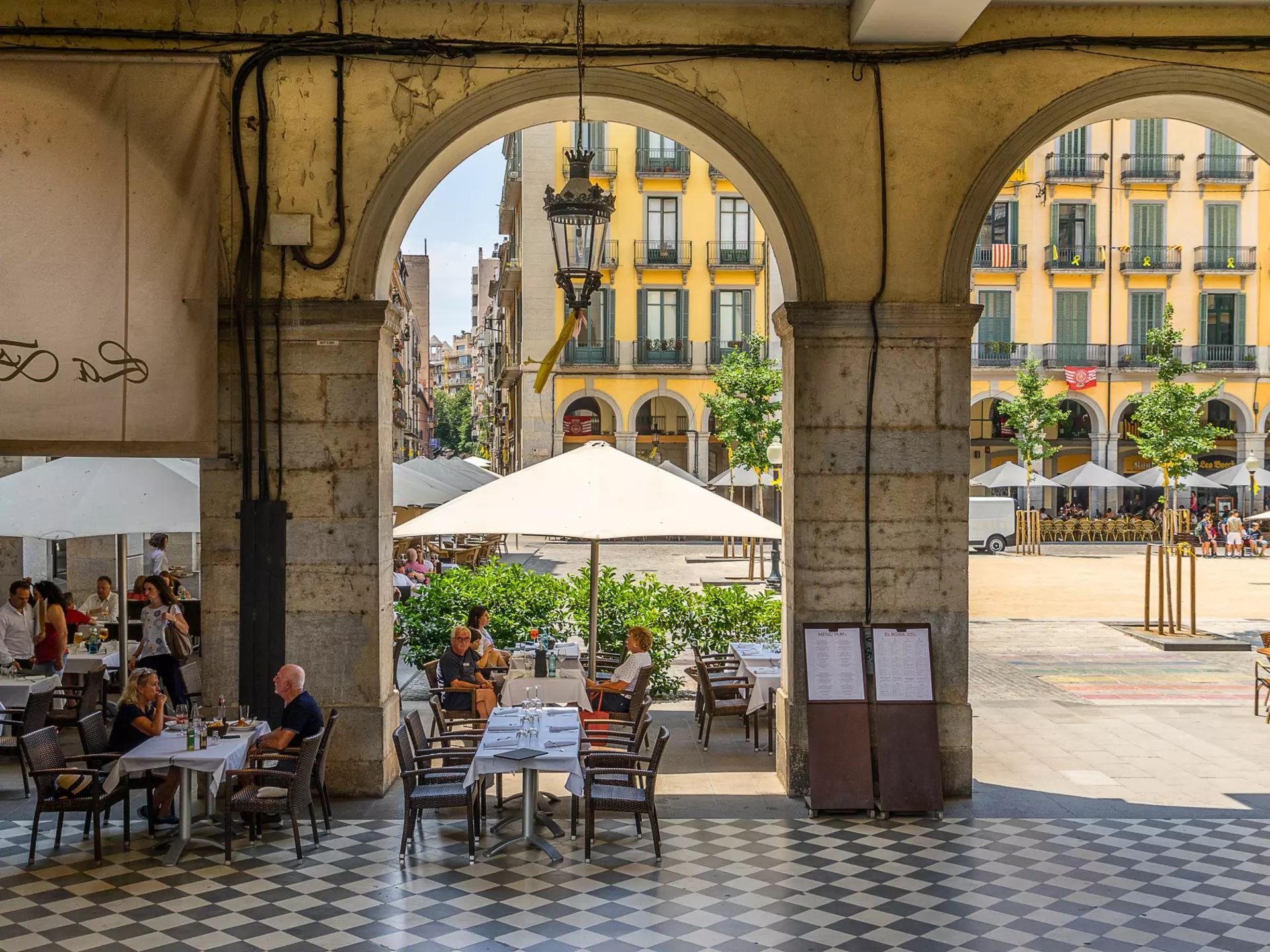 People sit at tables in a covered arcade next to a square in a city.