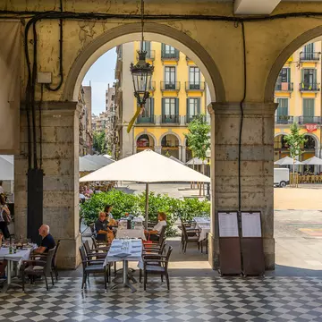 People sit at tables in a covered arcade next to a square in a city.
